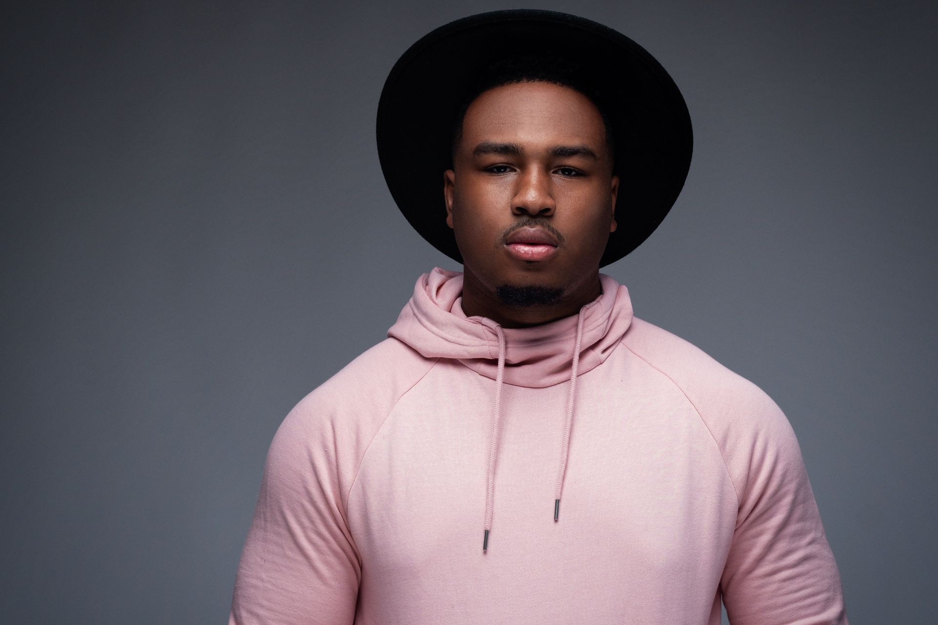 Man wearing a pink hoodie and black hat with a neutral expression against a gray background.